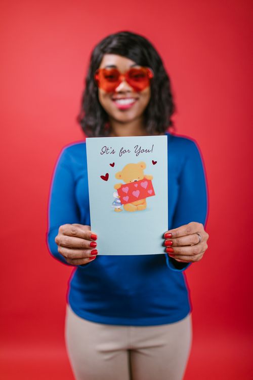 Free A cheerful woman with sunglasses holds a Valentine's card against a red background. Stock Photo