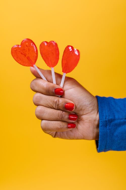 Free A hand with red nails holding three heart-shaped lollipops on a vibrant yellow background, perfect for Valentine themes. Stock Photo