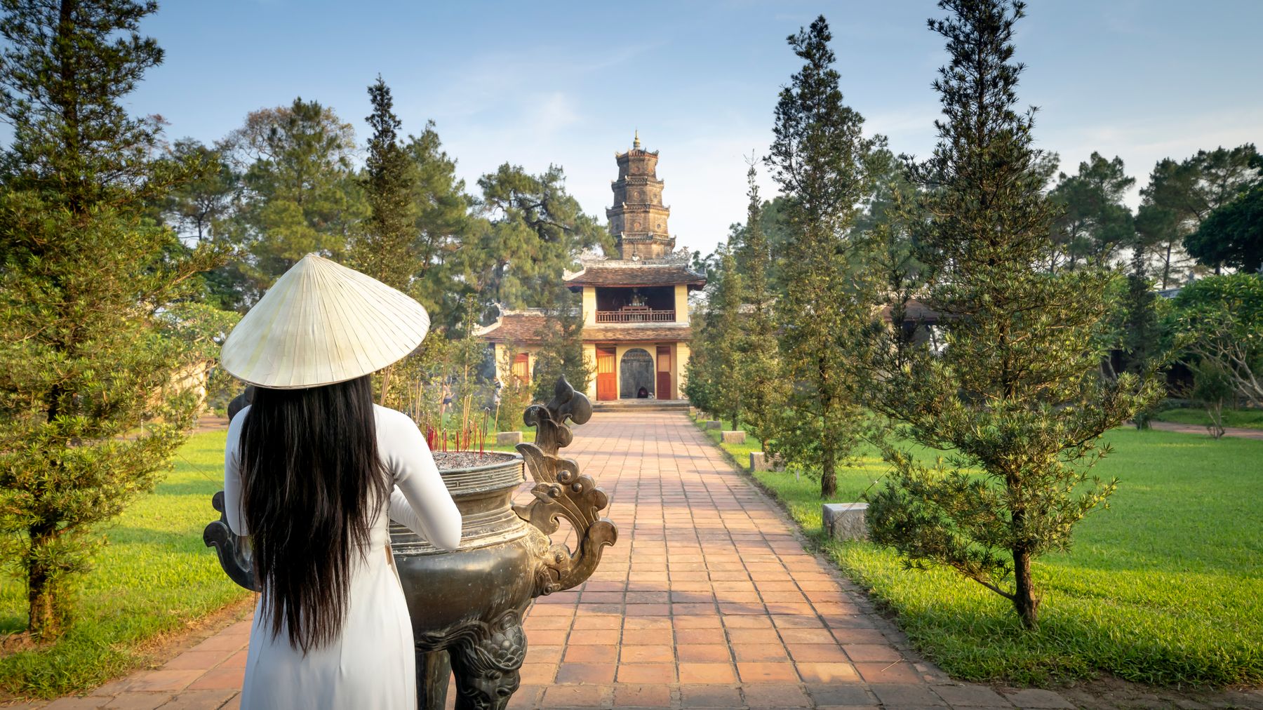 Back view of anonymous female in hat standing near green trees and pathway leading to old Pagoda Of The Celestial Lady