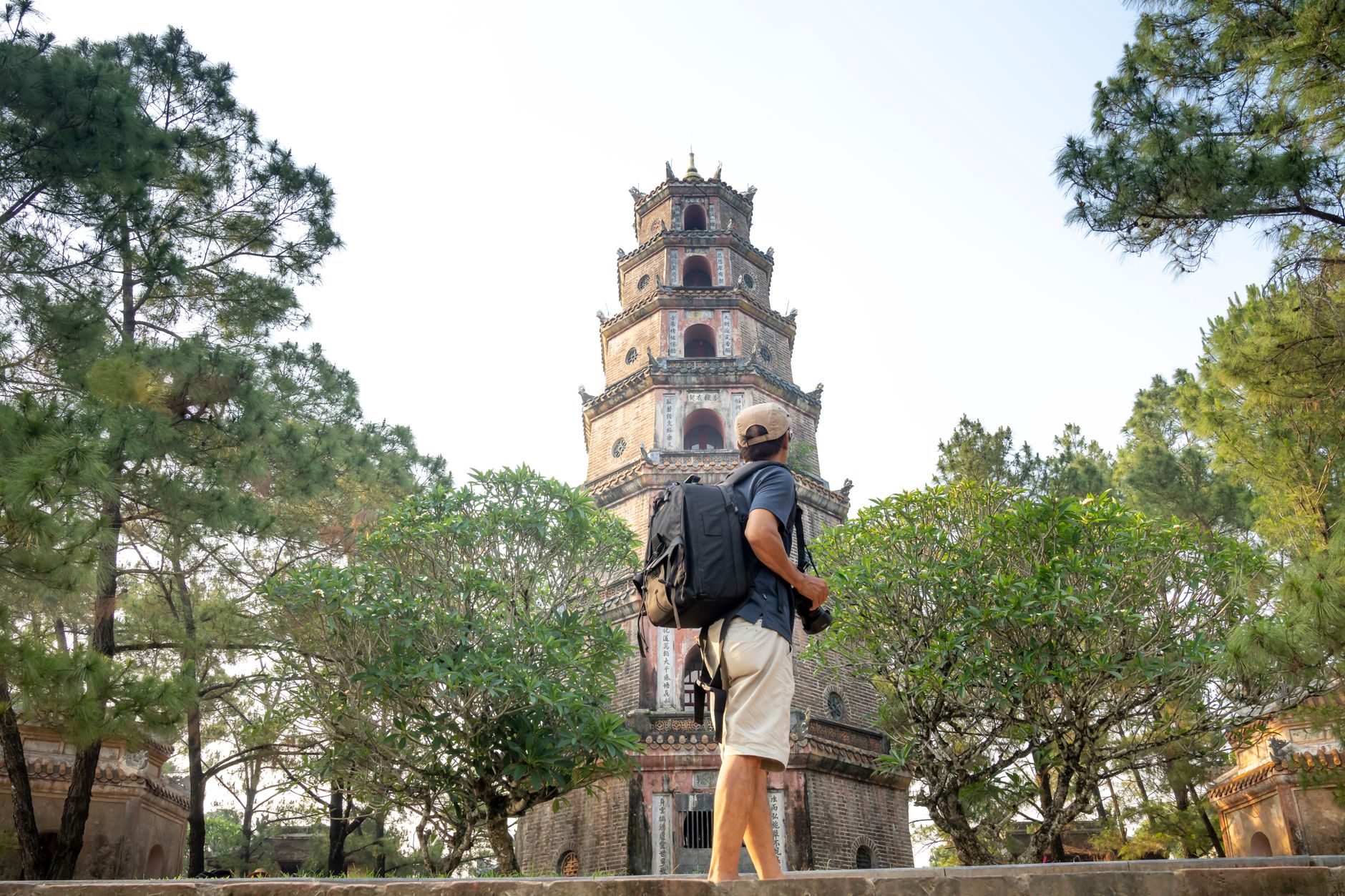 Side view of anonymous male tourist with backpack standing near green trees and old tower of temple during trip in Vietnam