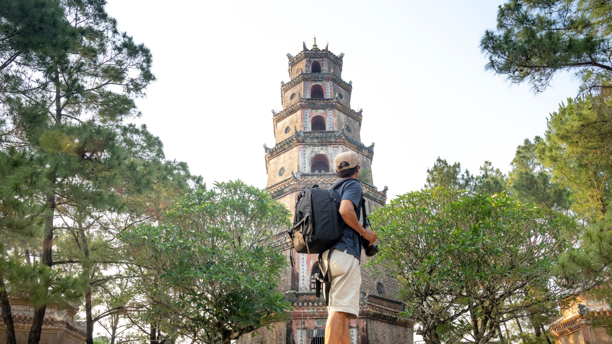 Side view of anonymous male tourist with backpack standing near green trees and old tower of temple during trip in Vietnam