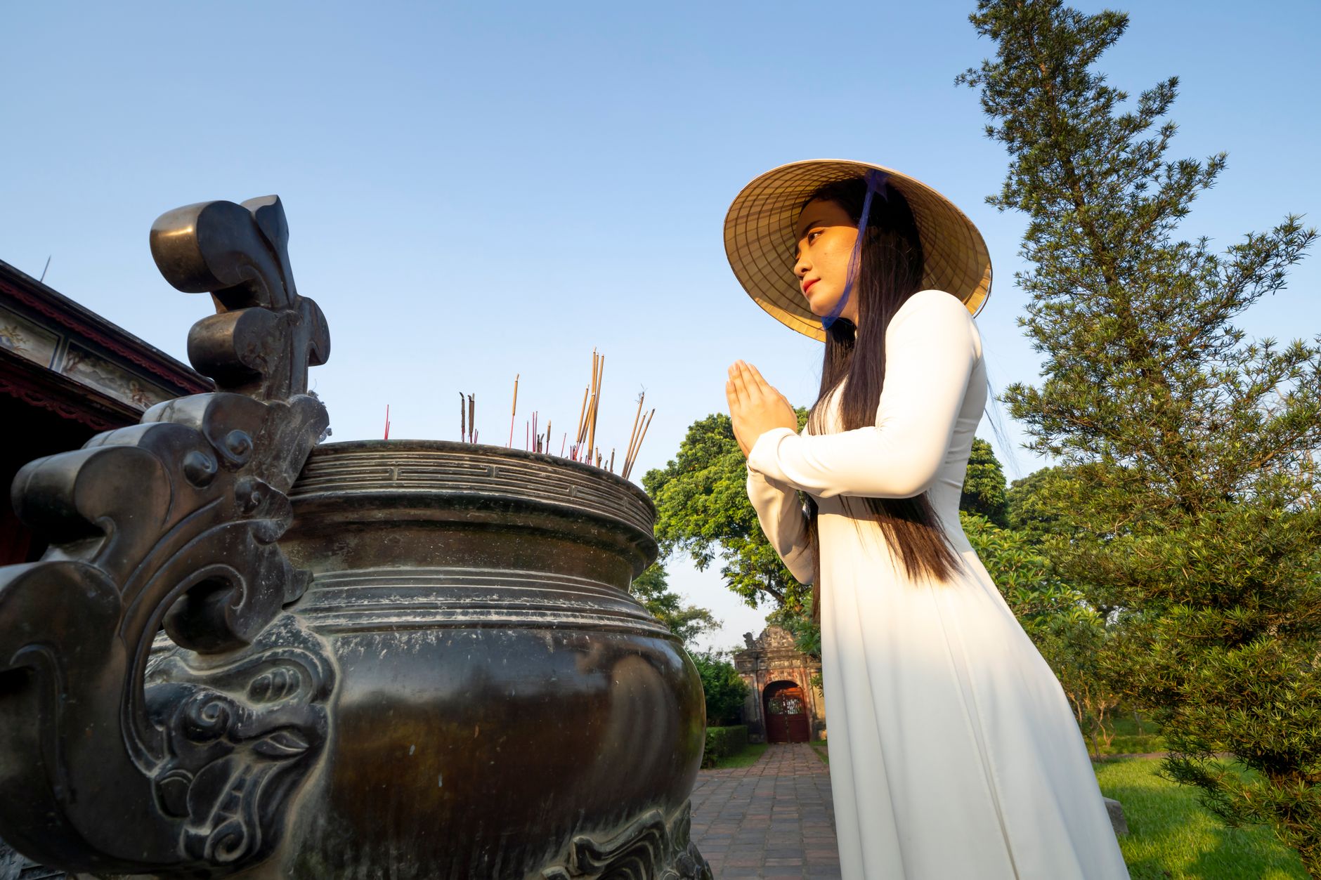 Side view of peaceful Asian female prayer with folded hands wearing hat standing near aged tripod on street in Vietnam