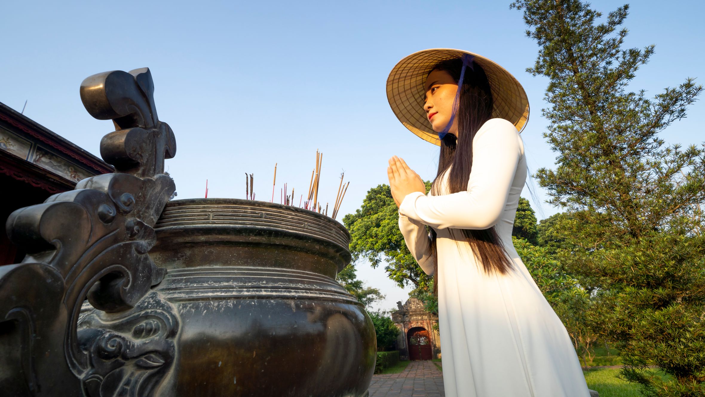 Side view of peaceful Asian female prayer with folded hands wearing hat standing near aged tripod on street in Vietnam