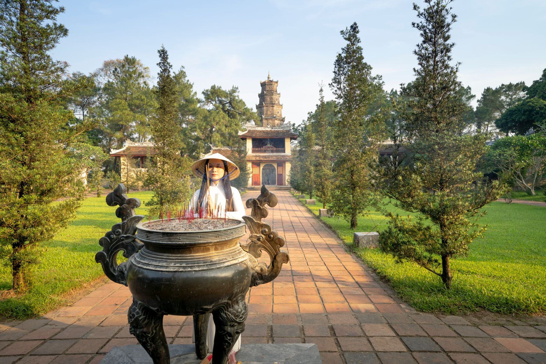 Asian female in hat standing on alley near traditional tripod against Pagoda Of The Celestial Lady near green trees in V