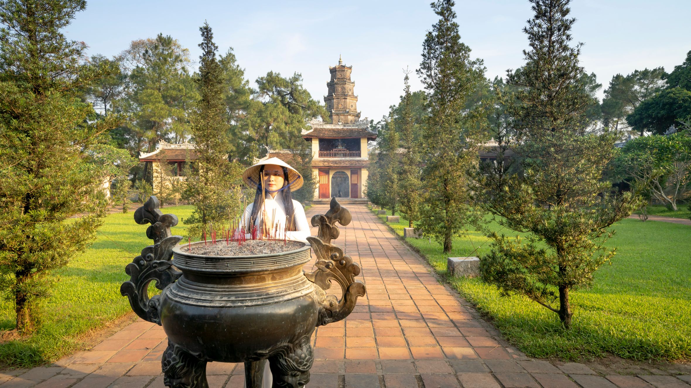 Asian female in hat standing on alley near traditional tripod against Pagoda Of The Celestial Lady near green trees in Vietnam