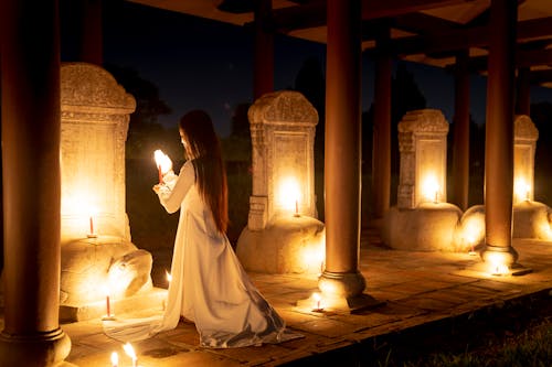 Full body back view of anonymous female with burning candles standing on knees near stone turtles while praying in dark time