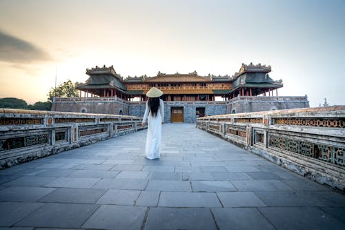Full body of anonymous woman with dark hair in long white dress walking on paved walkway near old oriental temple in daylight