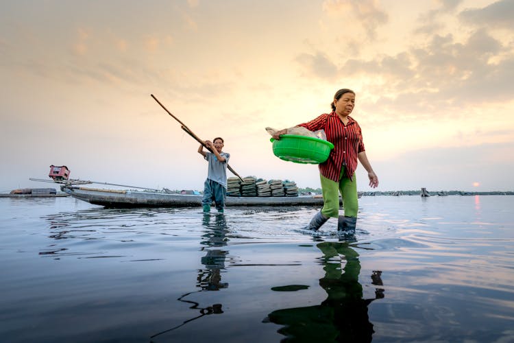 Asian Couple Walking In Water Near Boat With Fishing Tools