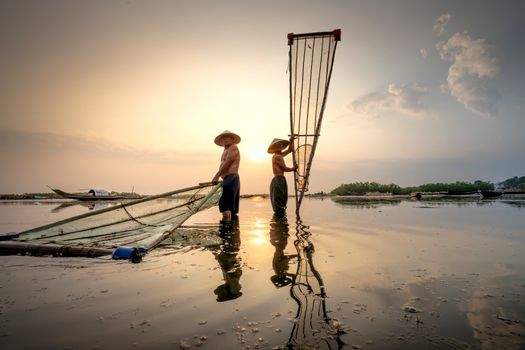 Asian fishermen in Vietnamese hats standing in river with fishing nets under cloudy sky in morning