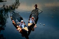 From above of ethnic female in dresses putting candle lanterns on lake water against anonymous male with oars in boats in twilight