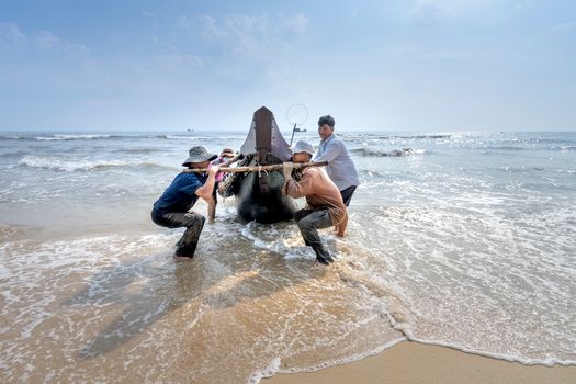 Side view of anonymous ethnic men dragging fishing boat on foamy ocean under cloudy blue sky in stormy weather