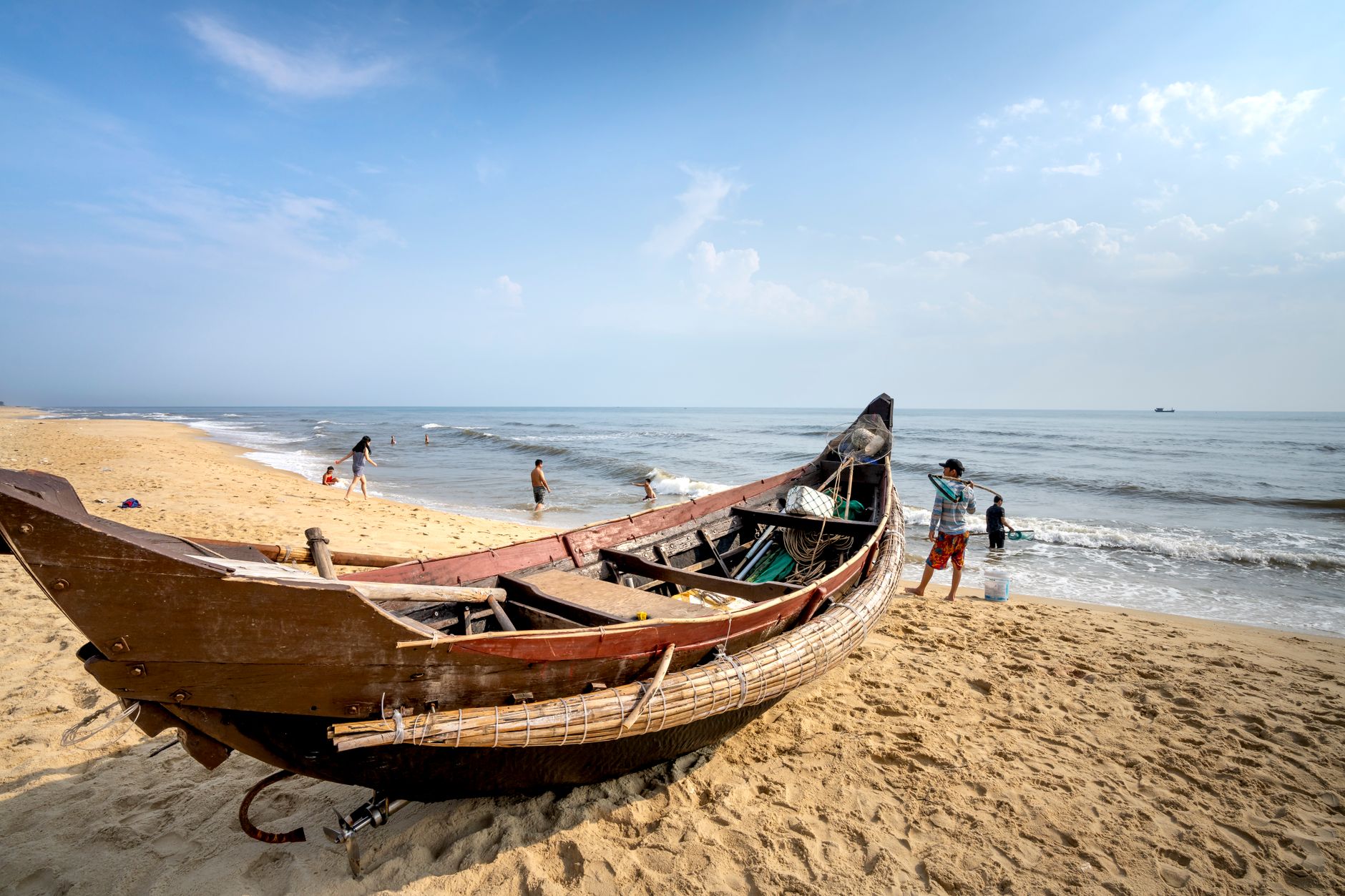 Anonymous tourists in wavy ocean against sandy coast with old oriental boat under cloudy blue sky in sunlight