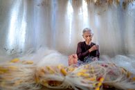 Elderly ethnic female with gray hair making fishing net with manual tool against shiny lights