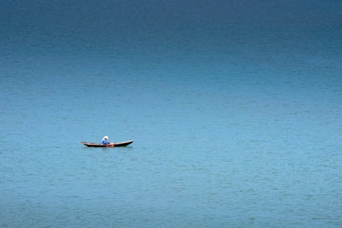 Side view of unrecognizable male fisher in conical hat sitting in fishing boat on rippled ocean in daylight