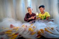 Focused elderly barefoot ethnic grandma with needle mending fishing net against boy with kerosene lamp