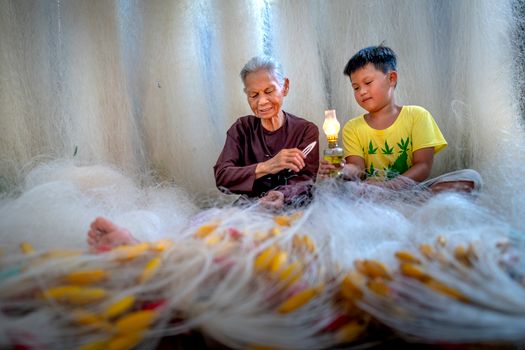 Focused elderly barefoot ethnic grandma with needle mending fishing net against boy with kerosene lamp