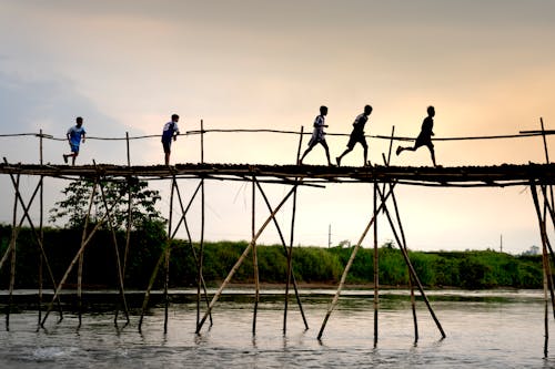 Unrecognizable active ethnic kids jogging on footbridge above lake while working out under sunset sky