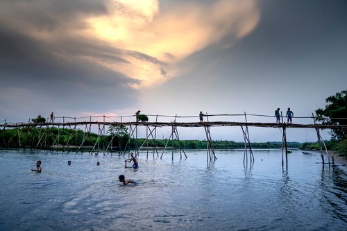 Free Anonymous ethnic men swimming in rippled lake while spending time under footbridge and cloudy sky in twilight Stock Photo