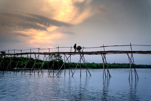 Free Anonymous male with wheelbarrow walking on bamboo bridge over rippled river under cloudy sky in twilight Stock Photo