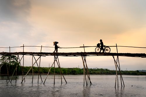Free Unrecognizable male silhouettes with bike on footbridge above rippled lake under cloudy sky in evening Stock Photo