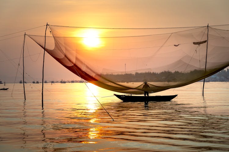 Anonymous Fisherman Standing In Boat Under Large Lift Net At Sunset