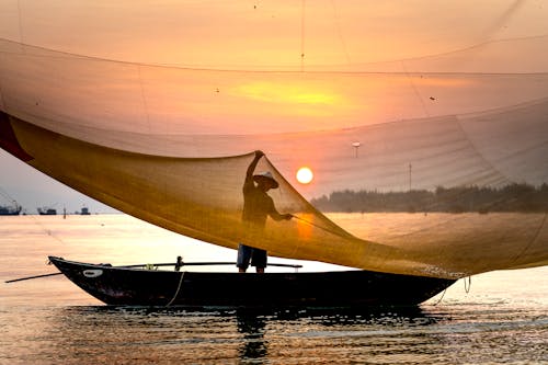 Anonymous male in hat throwing large fishing net while standing in boat floating on river at sunset in Vietnam