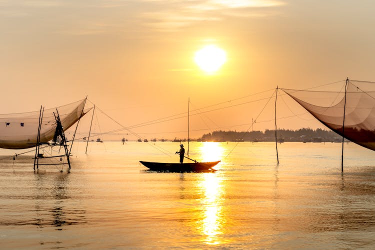 Unrecognizable Man Standing In Boat Between Fishing Nets At Sundown