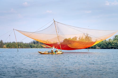 Free Anonymous local men in traditional fishing boat floating under large lift net against blue sky on sunny day in Hoi An Stock Photo