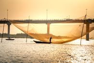 Silhouettes of unrecognizable people standing on boats while catching fish with large lift net near bridge over river at sunset in Vietnam