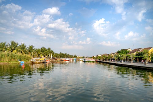 Picturesque scenery of colorful boats floating on calm river flowing between typical residential houses and lush tropical trees in Hoi An