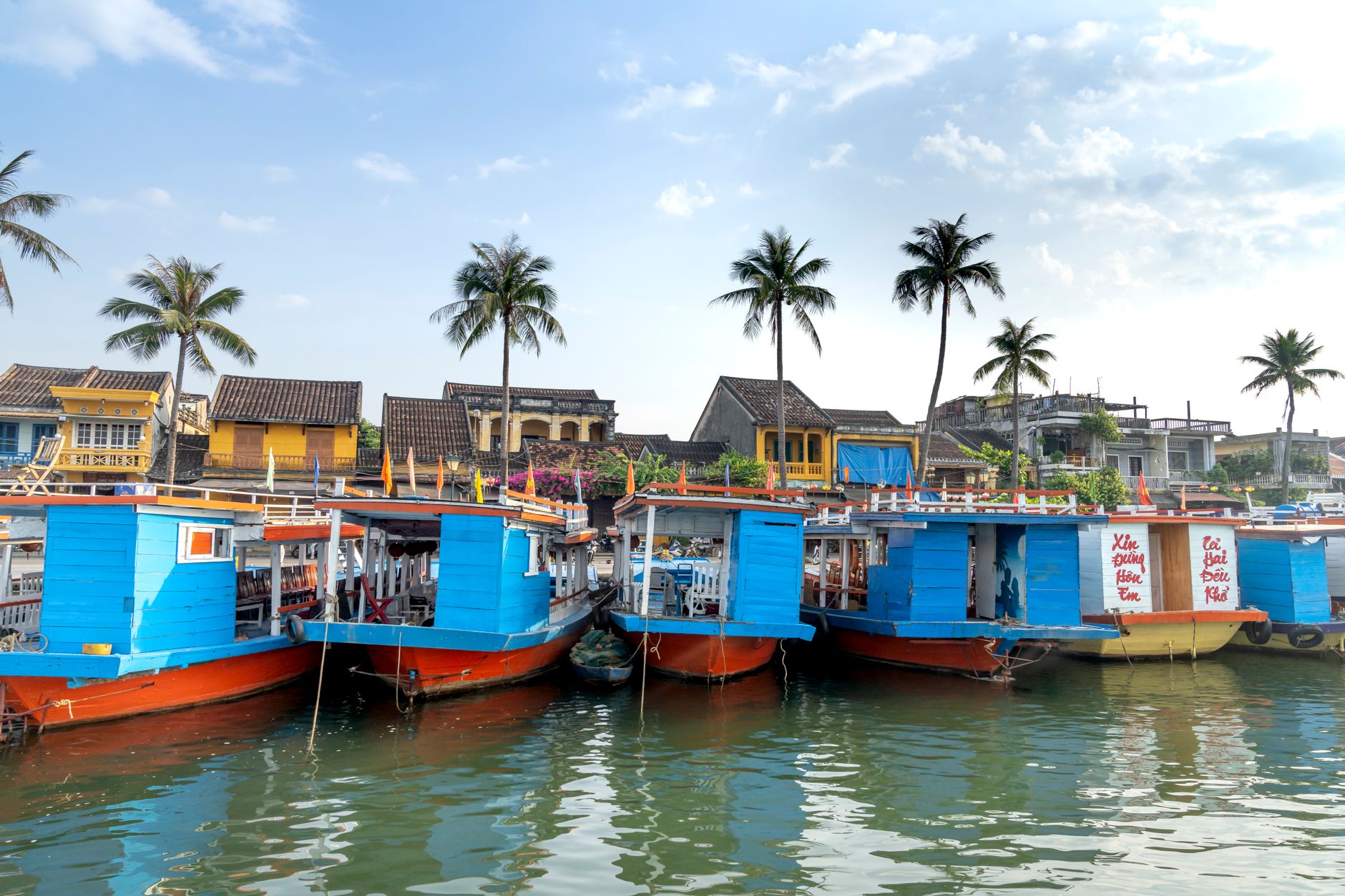 Excursion boats moored on river shore near typical residential cottages on sunny day in Hoi An