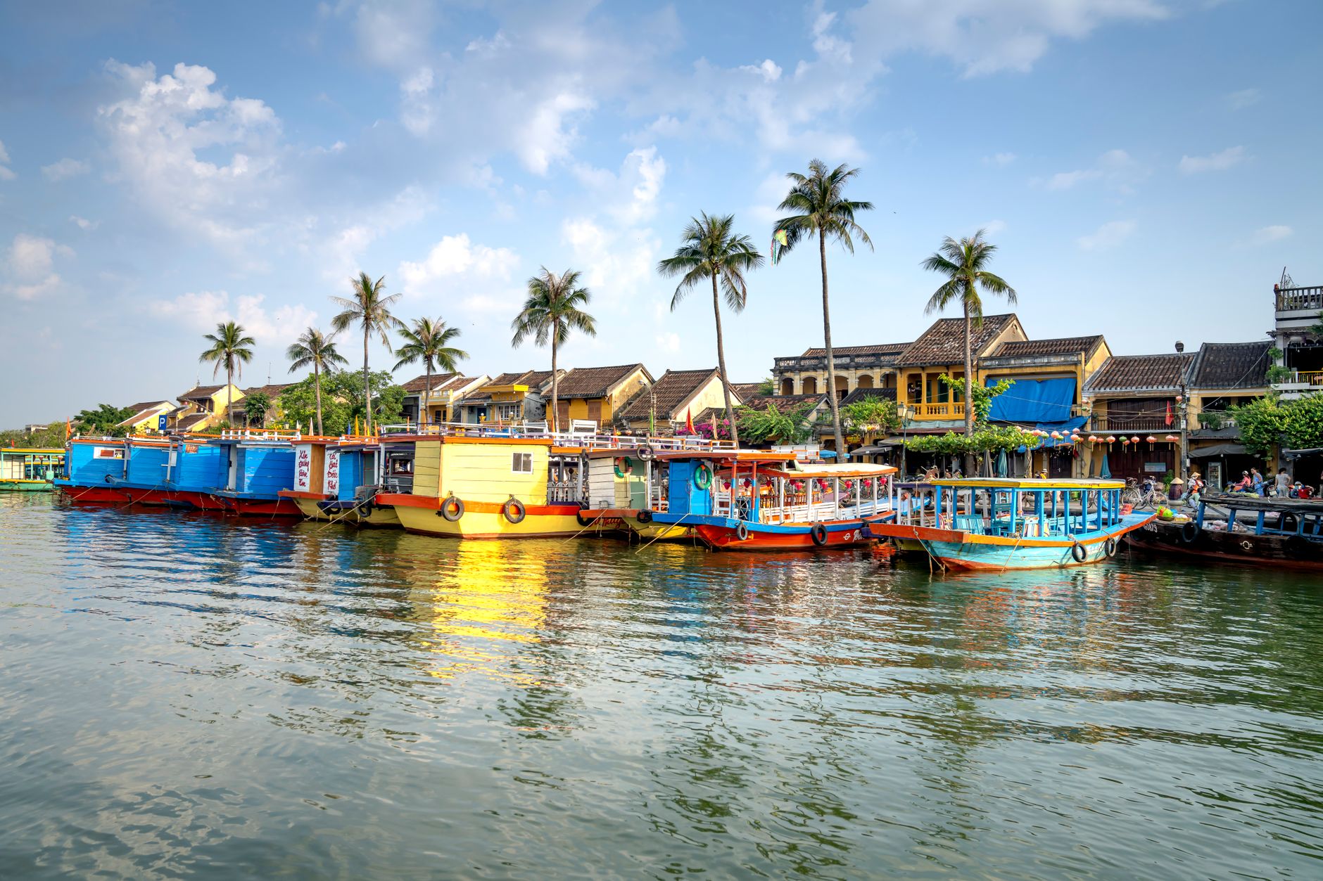Typical colorful boats moored on Thu Bon River near residential houses against blue sky in Hoi An
