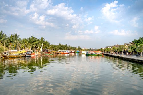 Free Scenic landscape of calm Thu Bon River with moored colorful boats flowing between lush palm trees and residential cottages in Hoi An on sunny day Stock Photo