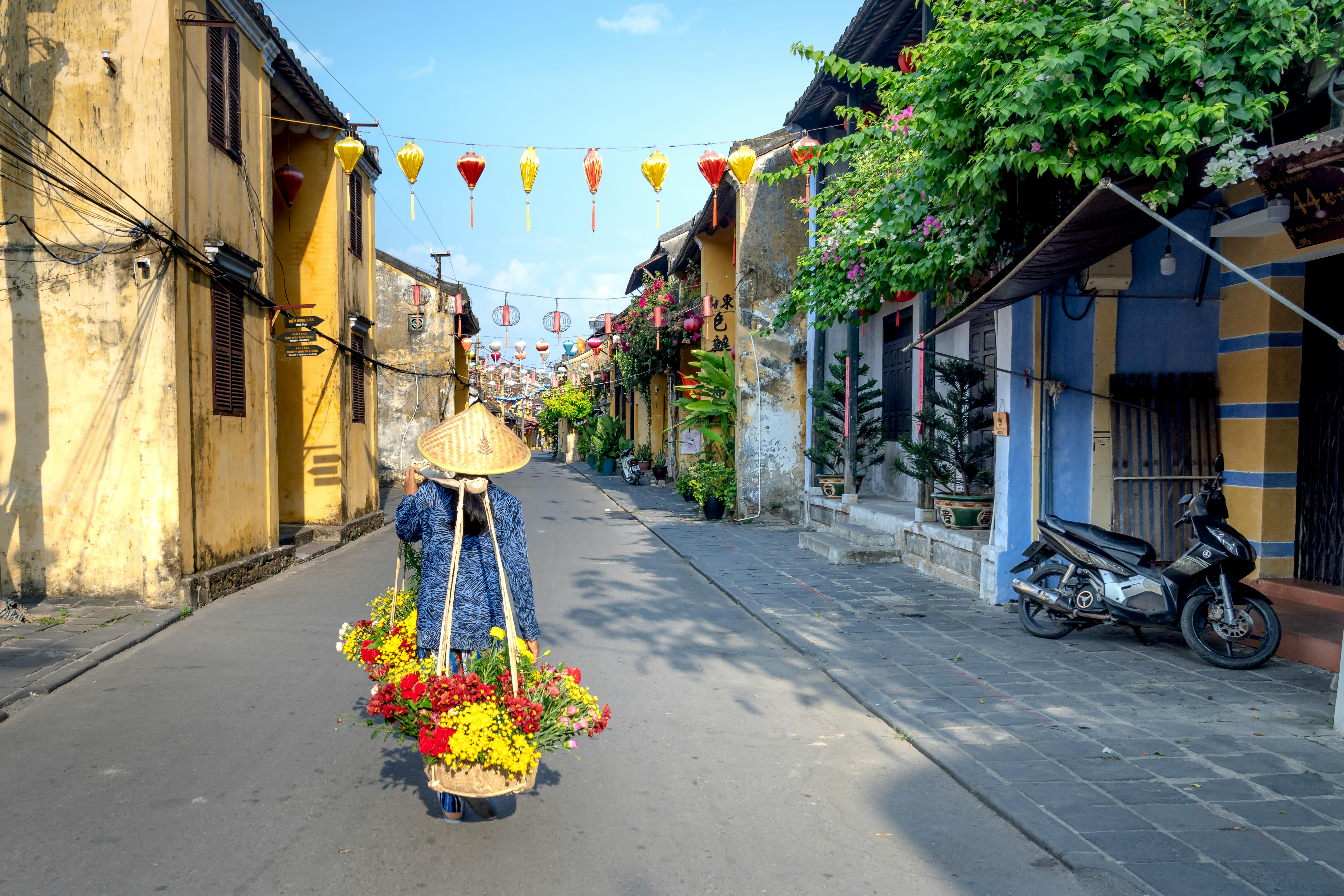 A woman with a conical hat carries flowers along a colorful street in Hoi An, Vietnam.