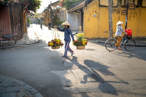 Side view of unrecognizable Vietnamese female seller in casual clothes and straw conical hat walking on Hoi An street with baskets of flowers near woman riding bicycle