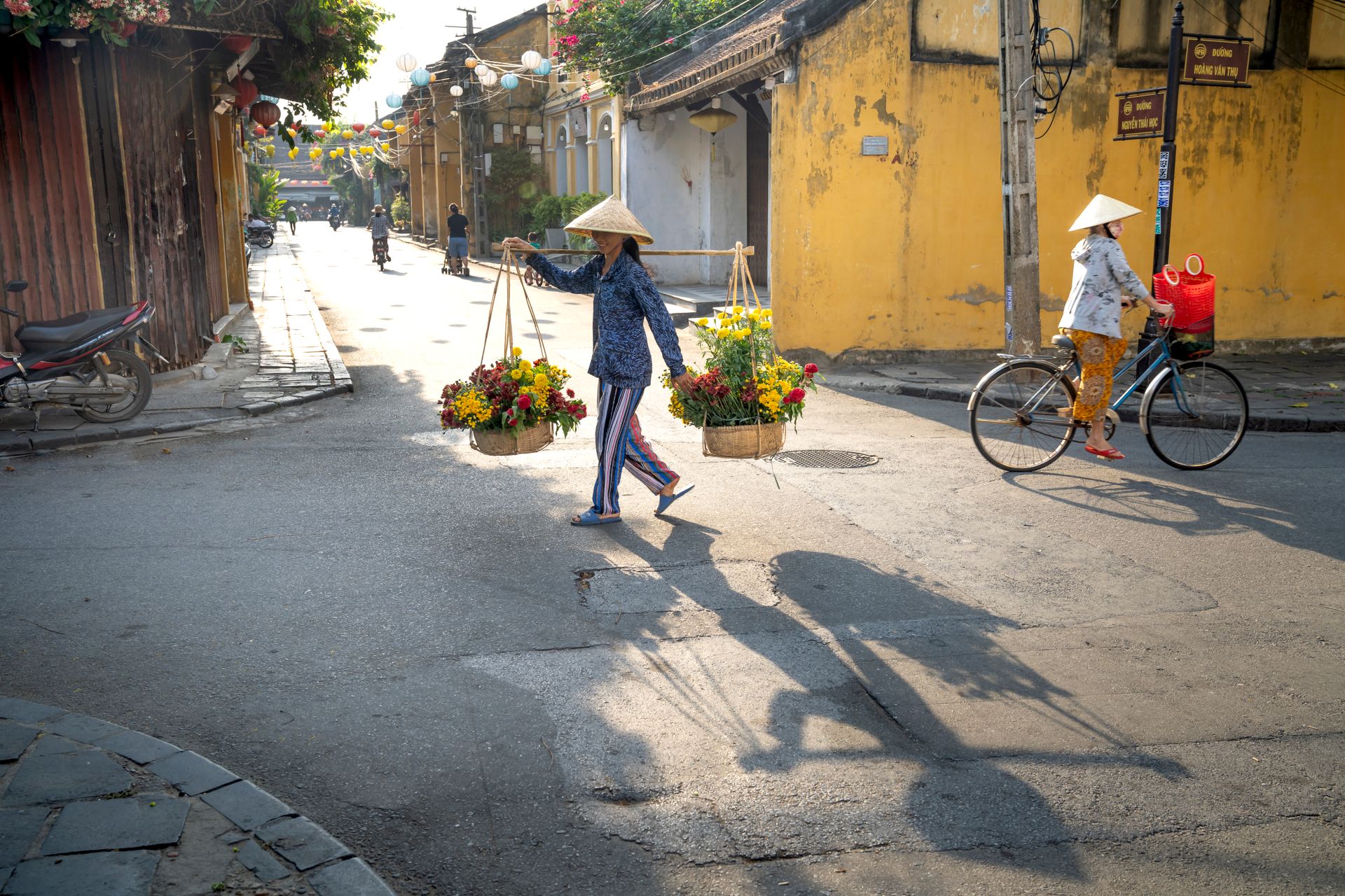 Side view of unrecognizable Vietnamese female seller in casual clothes and straw conical hat walking on Hoi An street with baskets of flowers near woman riding bicycle
