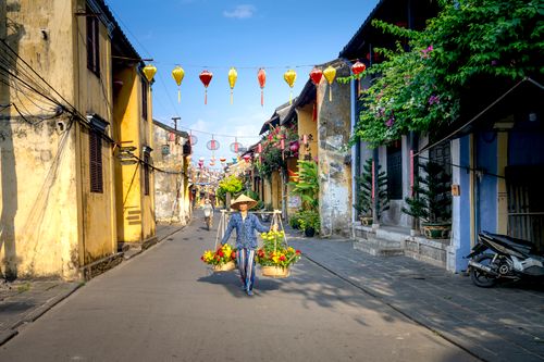 Free Full body of young Asian female flower seller in casual clothes and straw conical hat carrying baskets with bouquets while walking on street between residential houses of Hoi An Stock Photo