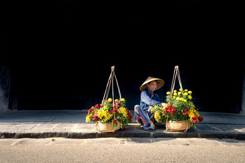 Free Full length of delighted young Vietnamese female street seller in casual clothes and traditional conical hat sitting on sidewalk near baskets with bunches of fresh aromatic flowers in town Stock Photo