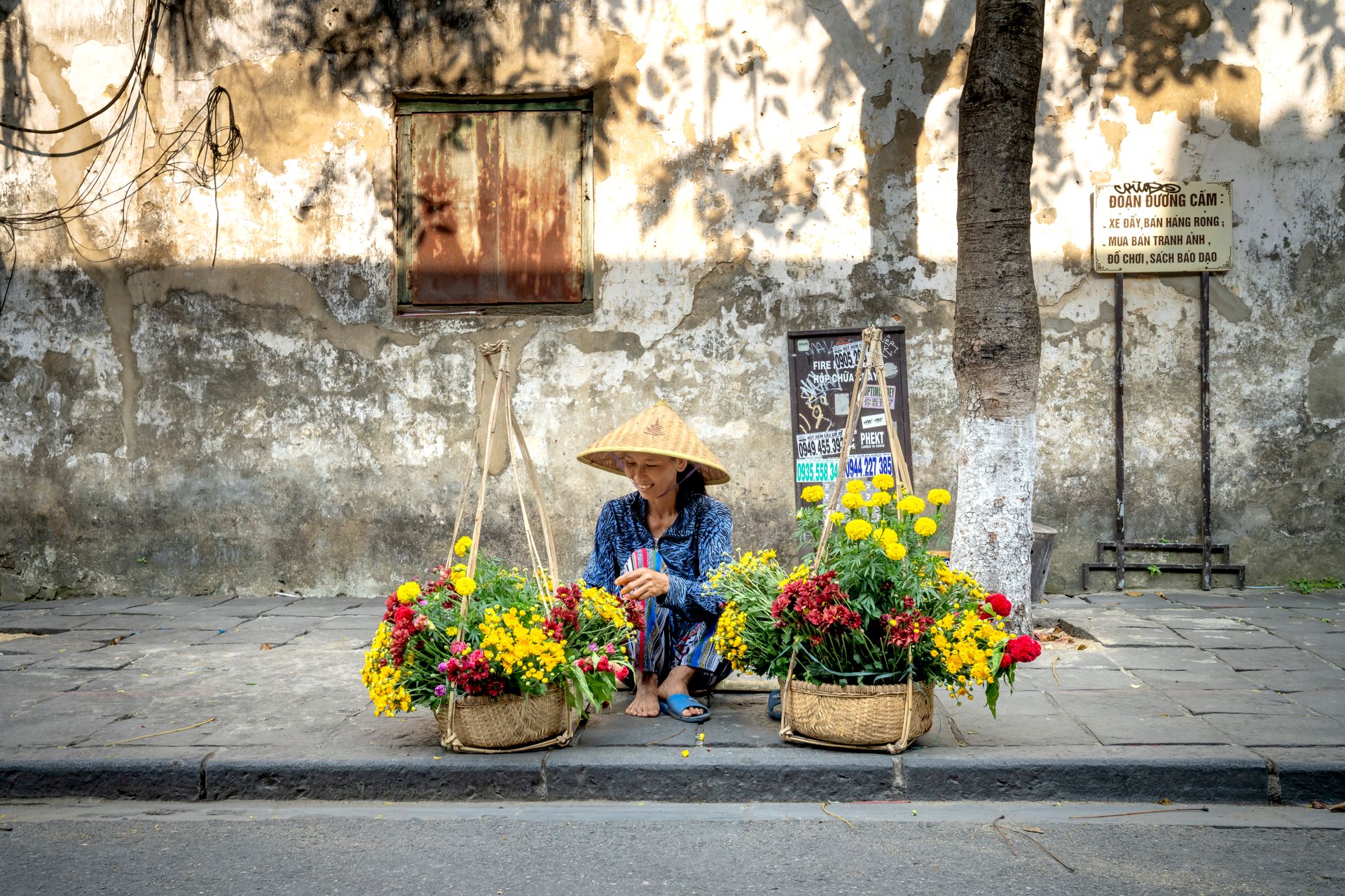 Side view of smiling young ethnic woman sitting on paves street in Hoi An near baskets with flowers while selling bouquets on sunny day