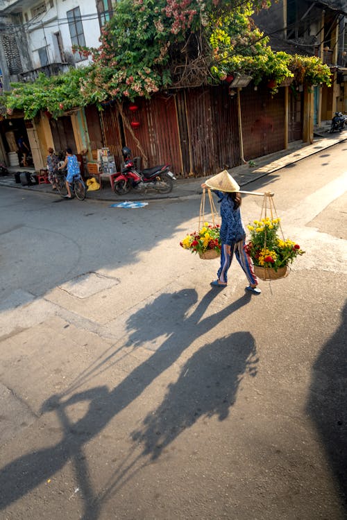 Anonymous Asian flower seller carrying heavy baskets while walking in town