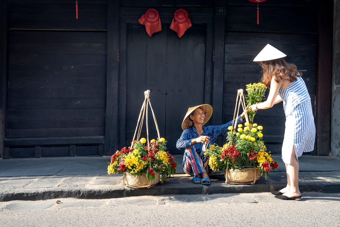Free Side view of young stylish lady in dress and conical hat buying bouquet of flowers from ethnic happy female seller sitting on street on sunny day in town Stock Photo