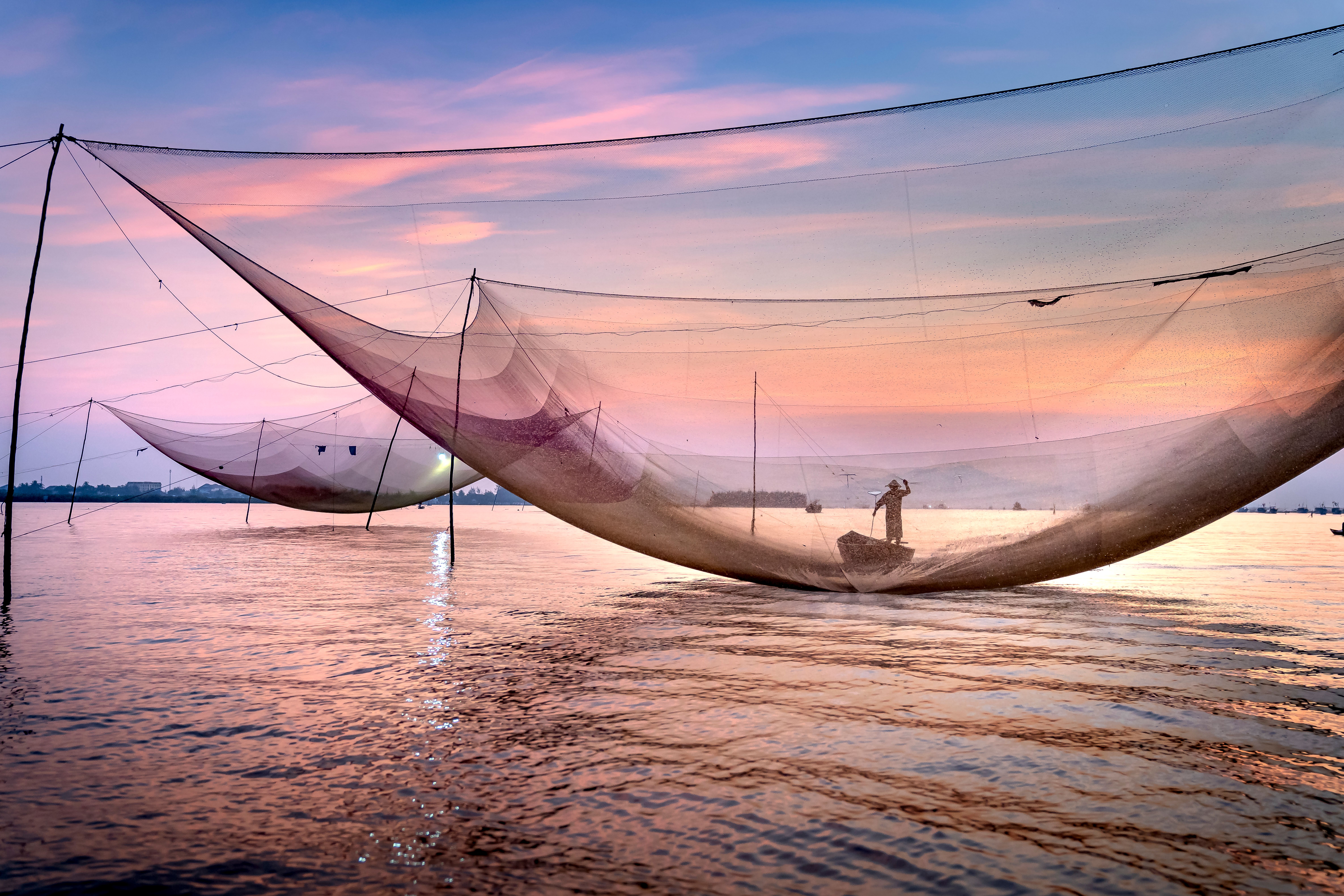 Distant unrecognizable male worker floating on vessel near big fishing nets on rippling sea against sundown sky on summer evening