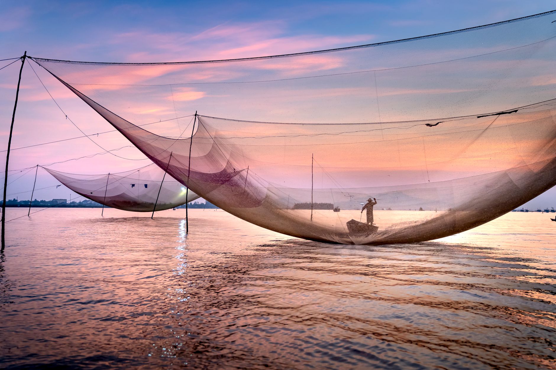 Distant unrecognizable male worker floating on vessel near big fishing nets on rippling sea against sundown sky on summer evening