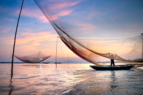 Silhouette of anonymous fisherman standing in boat under large lift net while catching fish against picturesque sunset sky in Hoi An