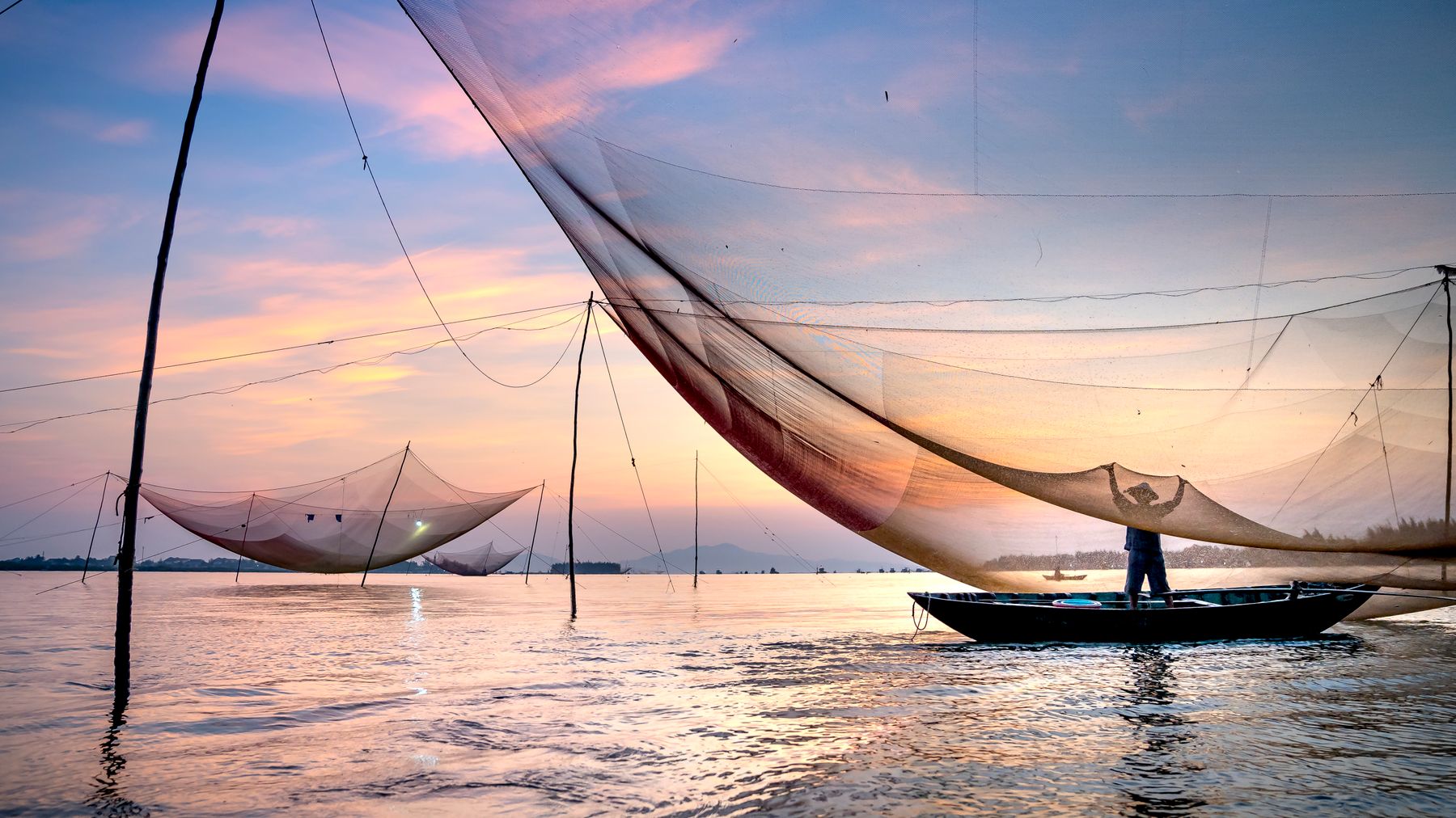 Silhouette of anonymous fisherman standing in boat under large lift net while catching fish against picturesque sunset s
