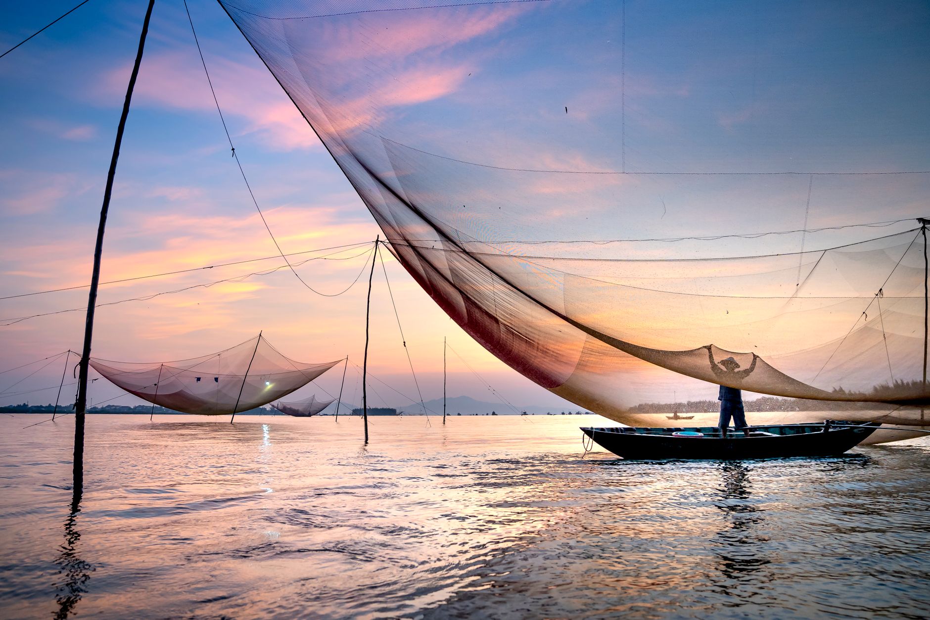 Silhouette eines anonymen Fischers, der in einem Boot unter einem großen Hebenetz steht und Fische fängt, vor einem malerischen Sonnenuntergang