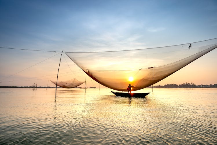 Silhouette Of Anonymous Man In Boat Catching Fish With Large Lift Net