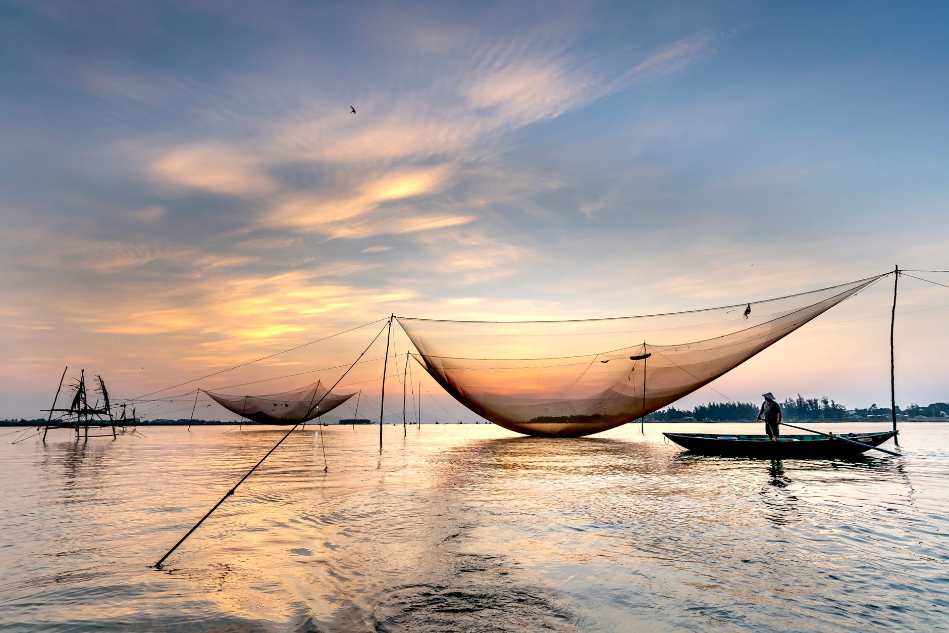 Anonymous male fisher rowing boat on river near large lift net against cloudy sunset sky in Vietnam