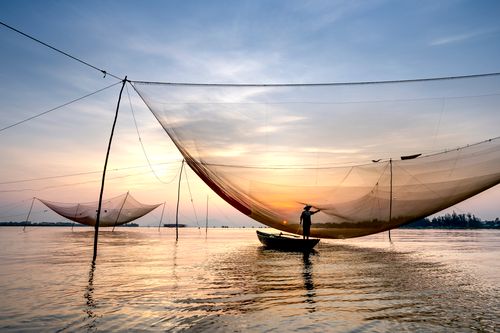 Free Back view of unrecognizable male angler standing in small boat on river while catching fish with large lift net at sunset in Vietnam Stock Photo