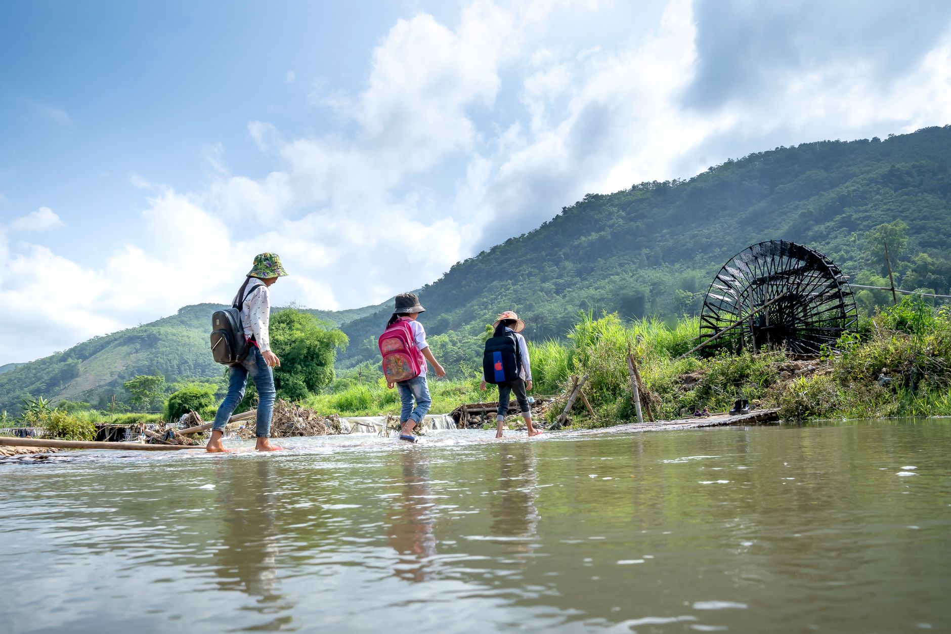 Low angle full body of barefoot girls with backpacks crossing river in hilly valley